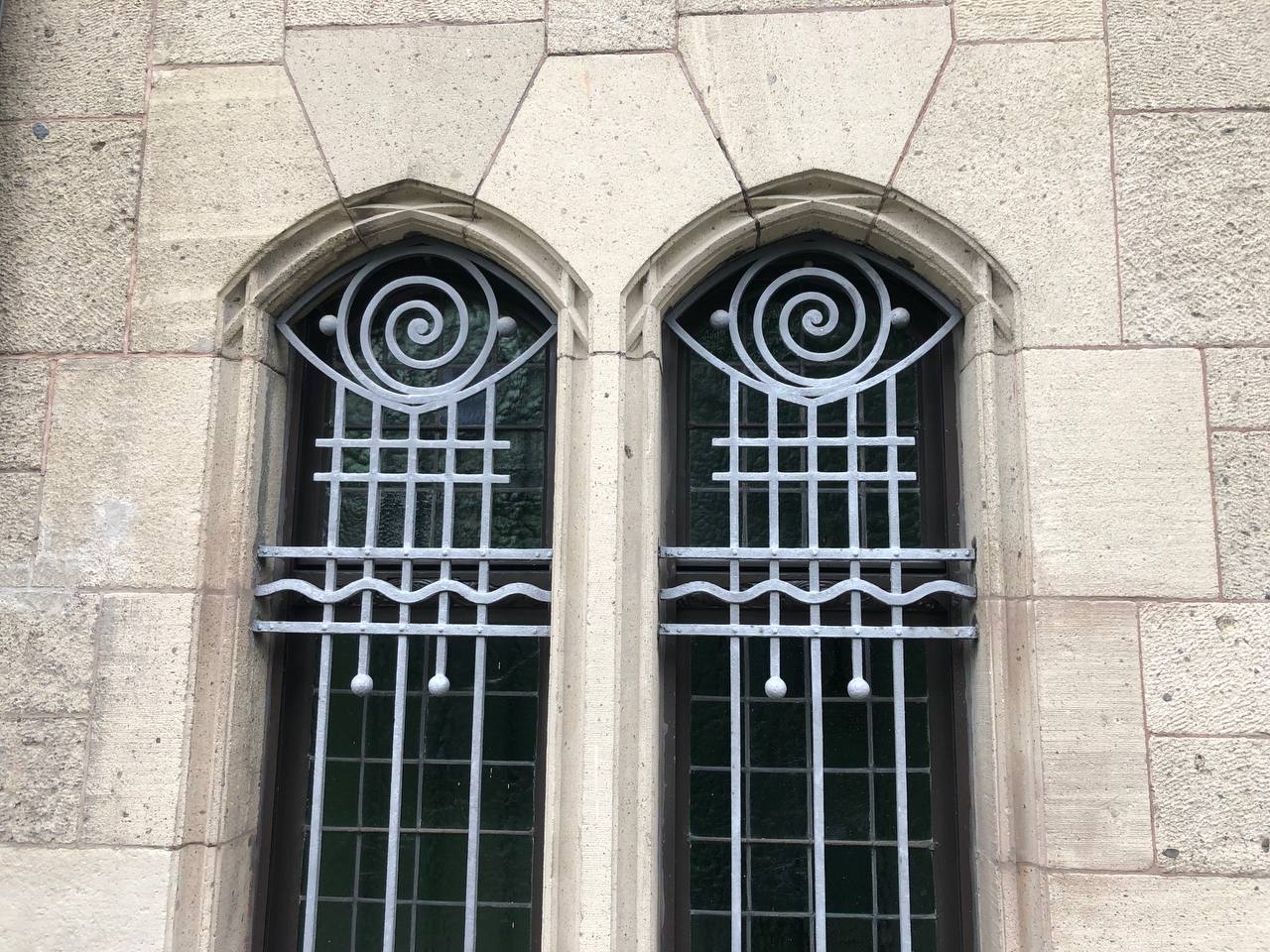 There are two arched windows set within a stone wall, each window adorned with an ornate iron scrollwork design. The scrollwork features a spiral pattern at the top, integrating security bars that extend down the windows. Both windows reflect this mirrored design, contributing to the architecture's symmetry and decorative appeal.