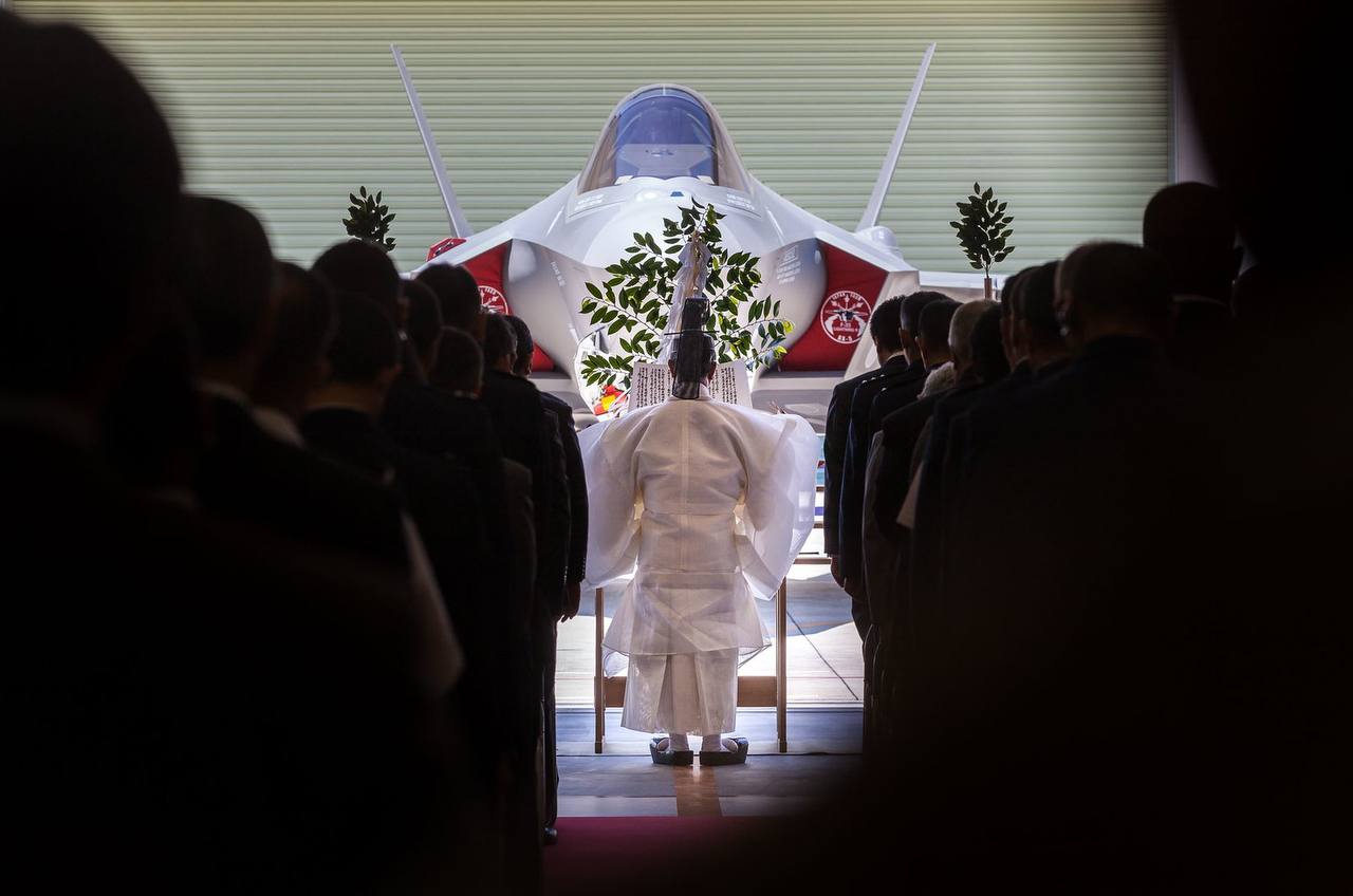 A group of people in formal attire are assembled in front of a jet aircraft, focusing their attention towards the center of the assembly, where an individual in white traditional ceremonial robes is standing, with their back to the camera. The aircraft visible features stealth characteristics and a twin-tail design, suggesting it is a modern military fighter jet.