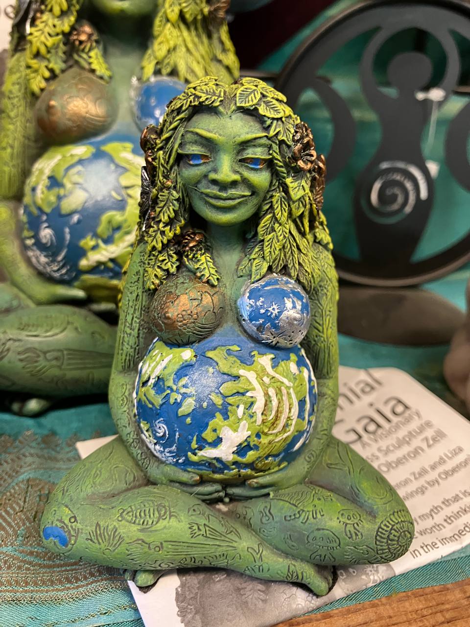 A figurine depicts a maternal representation of nature, personified as a female entity with green leafy hair and whispers of plants and leaves making up its form. With a content smile, she holds a spherical depiction of the Earth close to her chest, evoking a sense of protection and nurturing.