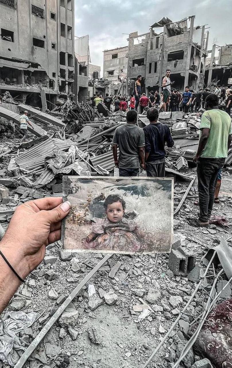 A hand holds a photograph of a child amidst a backdrop of destruction with collapsed buildings and debris. People stand and sit on the piles of rubble, and works seem ongoing with many observers around. The color in the scene focuses on the photograph, which also appears dirty and worn, suggesting a personal memory amidst the chaos of the environment.