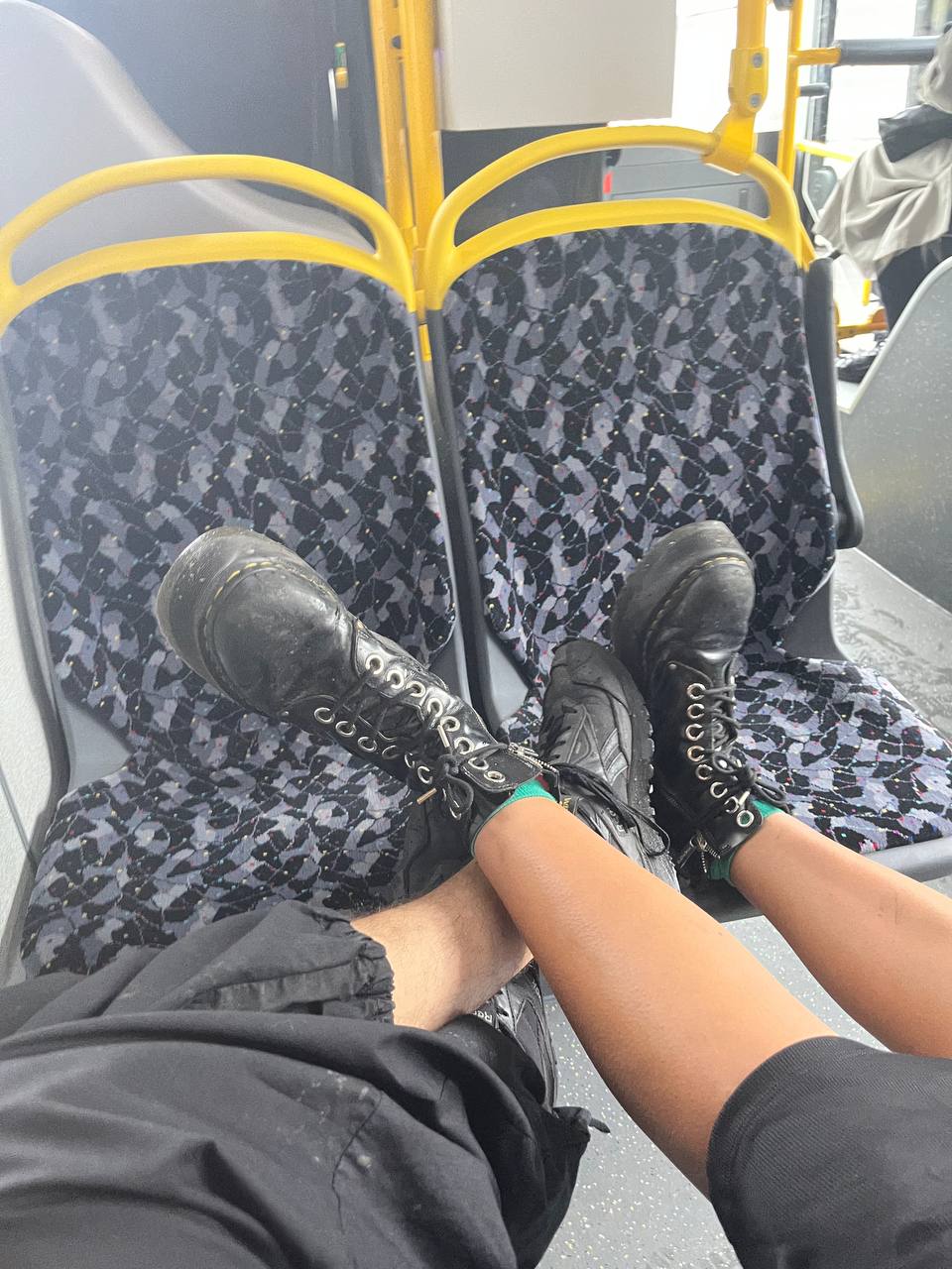 There are two legs crossed at the ankles resting on a bus seat. The person is wearing black, high top boots with laces, black pants, and sporting what appears to be a smartphone or other partly visible items nestled beside the seat.