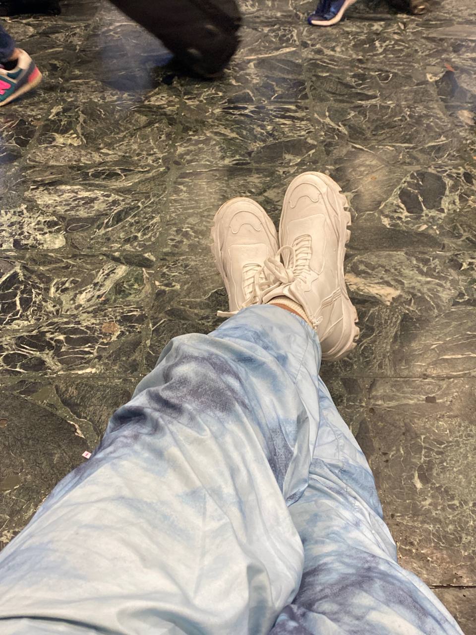 A pair of crossed legs clad in blue tie-dyed jeans and wearing chunky off-white sneakers are prominently displayed, while in the background, the dark marble floor with various black and green hues where blurry movements suggests the restlessness of possibly an airport or a similarly busy venue with feet wearing other colorful sneakers passing by.