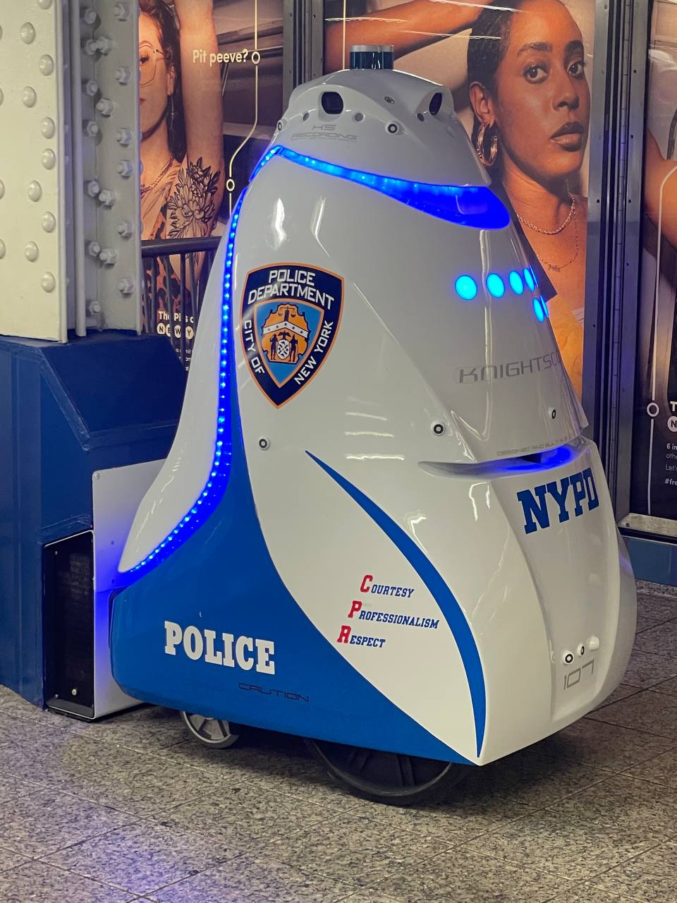 A robotic device with a blue and white design, labeled with "POLICE," the NYPD (New York Police Department) emblem, and the words "Courtesy, Professionalism, Respect" appears prominent in the forefront. It's equipped with blue lights akin to those on police cars. Advertisements featuring images of people are partially visible in the background.