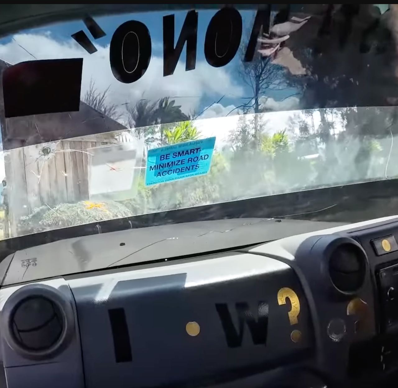 Looking out from the interior of a vehicle, the view is partially obscured by multiple reflections and possibly dirt on the windshield. Sunlight casts reflected images and glare onto the windshield, highly prominent on the left.