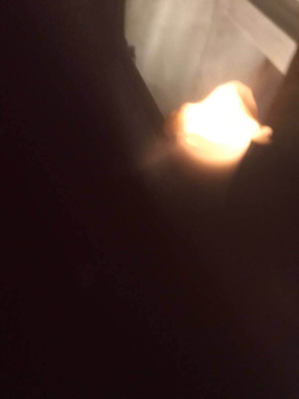 The focus seems to be on a blurred bright spot which could be a light source, possibly a flame, against a largely dark background with little to no discernible detail. The quality of the photo is poor and doesn't allow for a clear understanding of the environment in which this light source is present.