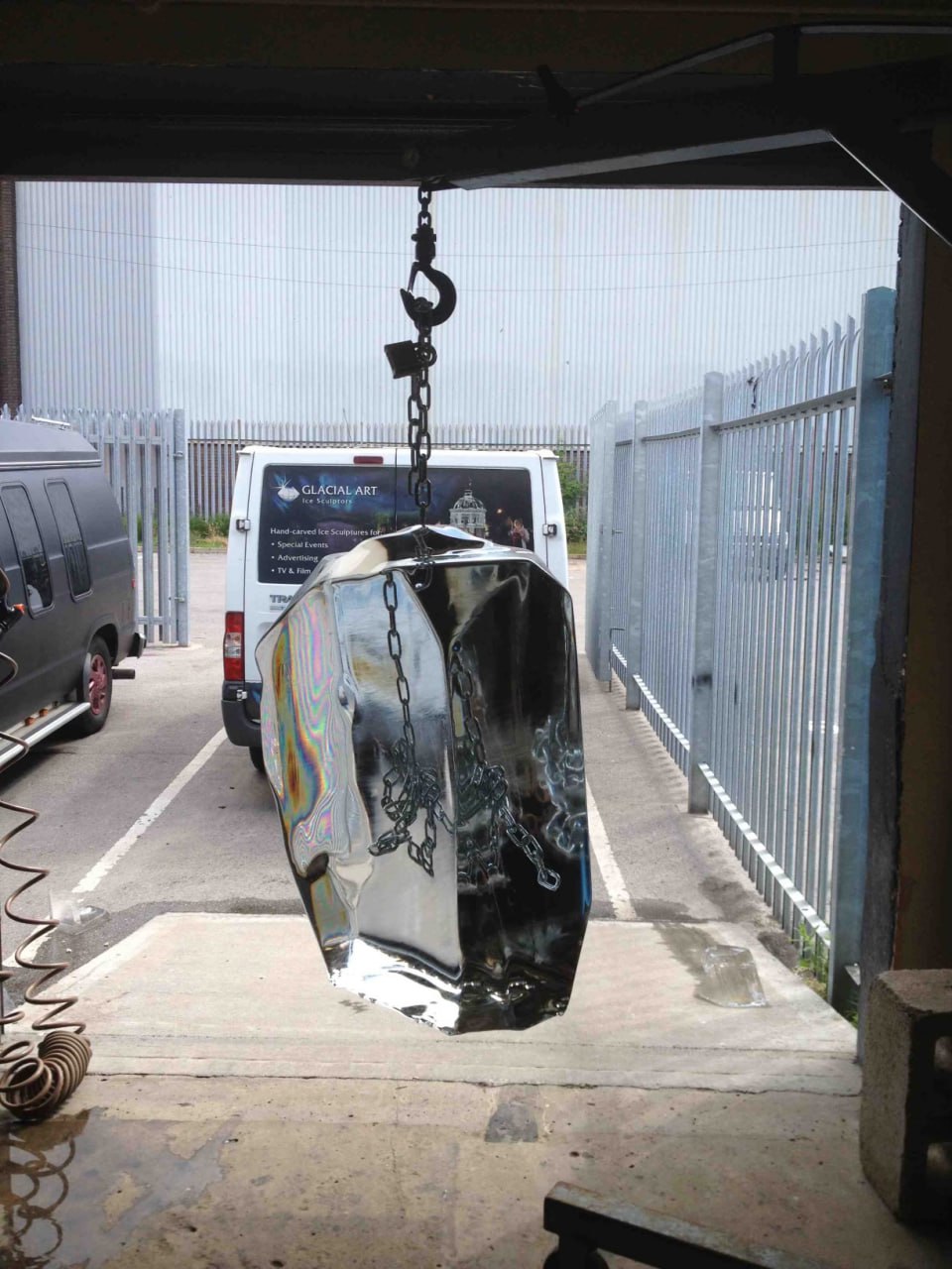 A large, clear ice sculpture dangles from a chain attached to a metal hook, likely inside a workshop or delivery area. Good lighting accentuates the sculpture's transparency and smooth edges. In the background, a van featuring the word "GLACIAL ART" is visible, suggesting that the icy sculpture may have been produced or is being delivered by a company specializing in crafted ice pieces.