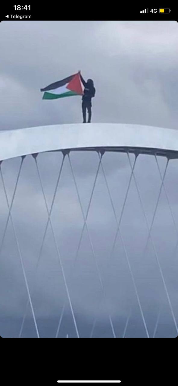 A person stands at the top of an arched structure, holding a flag with a horizontal tricolor design and a triangle juxtaposing the bands at the hoist.