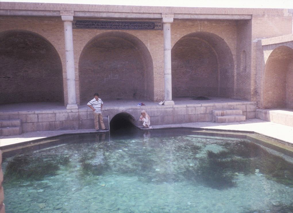 A brick structure with arches surrounds a clear, circular body of water which appears to be a traditional pool or pond. Two individuals are leaning by the edge on the left, and one person is seen near a small passageway or tunnel that leads under the structure. The focus reflects a serene atmosphere most likely a part of a historical or cultural site.