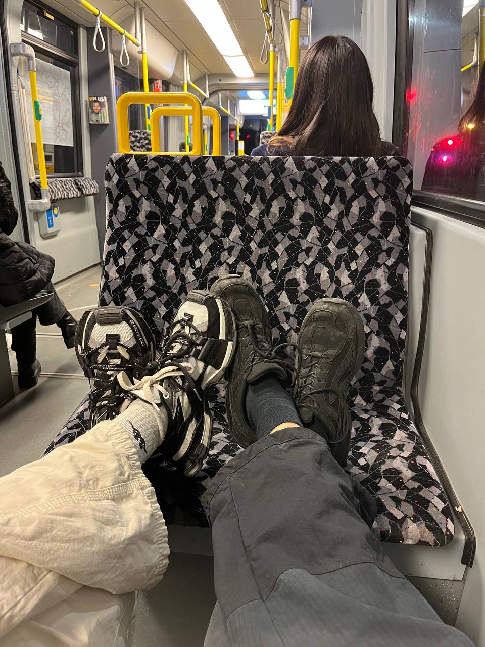 Two pairs of legs and feet resting on the seat of a public transit vehicle, with shoes on. One set wears off-white cargo pants with white socks, paired with black and white athletic shoes. The other is clad in dark trousers with dark socks, along with solid black rugged-looking shoes. The vehicle interior is also visible, featuring patterned seat fabric and a window reflecting light from outside.