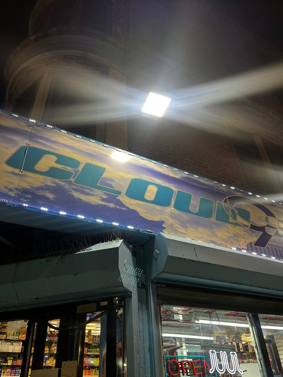 The storefront of a shop illuminated by bright lights at night is seen. A blue lit sign with the word "CLOUD9" in capital letters, likely a brand or shop name. Along the edge of the roof, directly beneath the sign, there is a blue light strip and spikes installed to deter birds. There is a strong light source near the ceiling, creating a lens flare effect in the visibility.