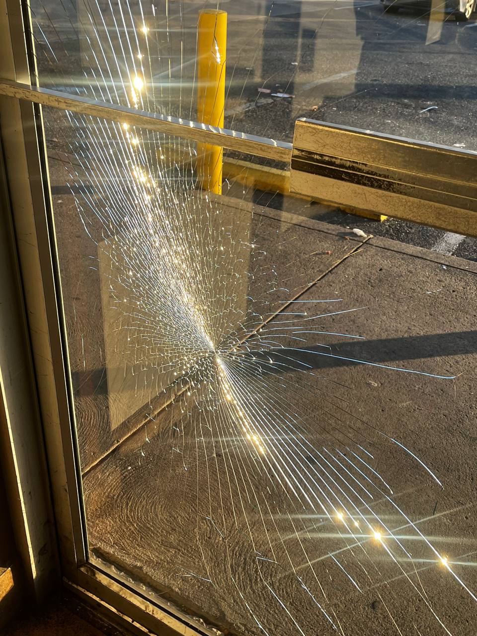 Visible is a shattered glass door or window with what appears to be sunlight beaming through it, casting a bright glow at some of the break points. The shattered rippled pattern suggests the glass is likely safety glass, designed to hold together even when broken.