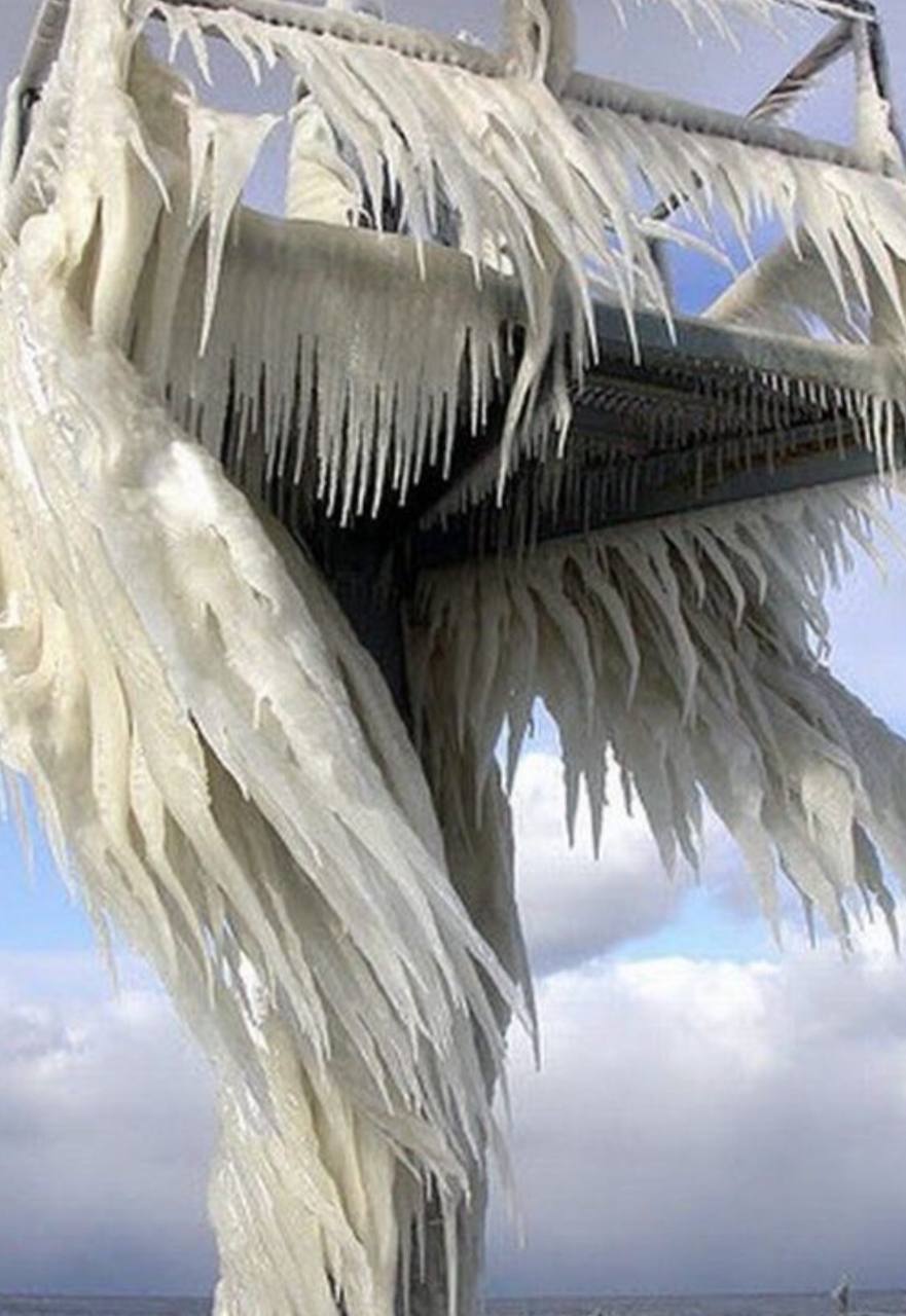 Icicles have formed on a vertical structure, creating an almost feather-like appearance. The skew and directionality suggest that wind played a role while the ice was forming. The structural components appear to include metal beams, some of which run horizontally, while others create a box-shaped framework, all thickly coated with ice.