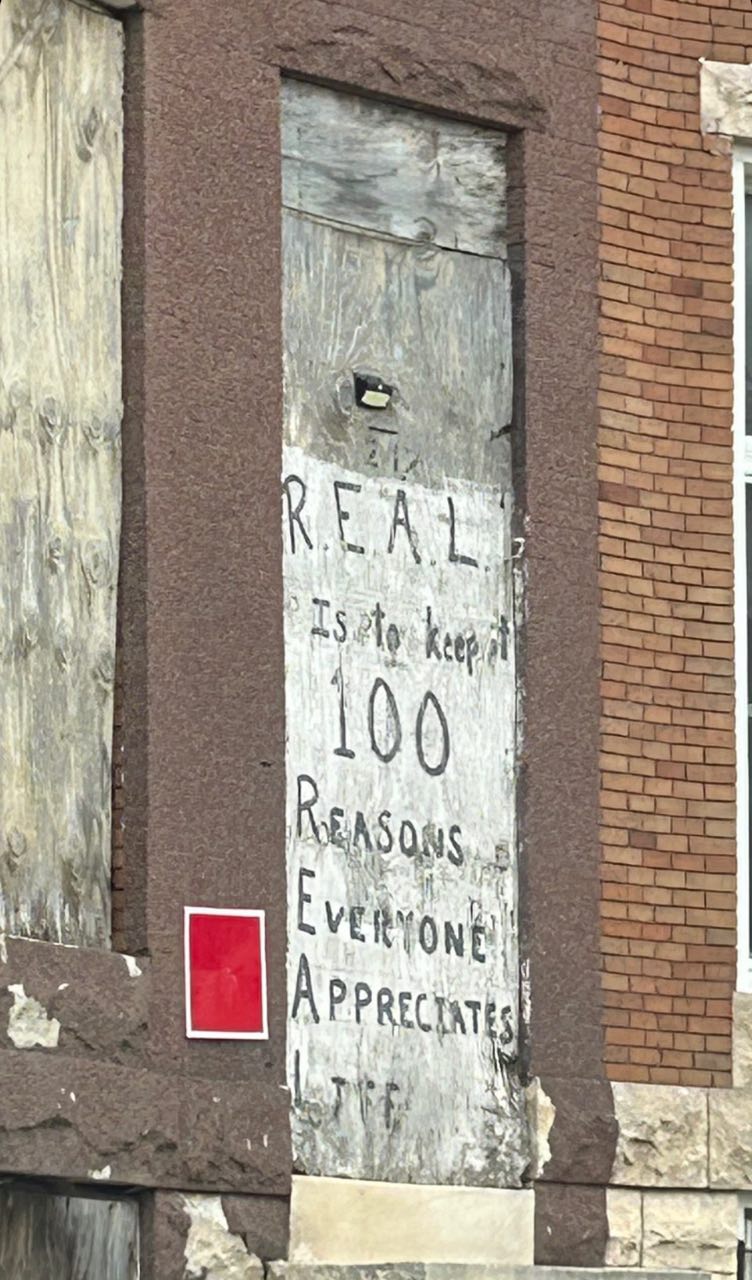 Weathered wooden door boarded up, graffiti in black paint forming an acronym "REAL," each letter beginning a new line—"R.E.A.L. Is to keep 100 Reasons EveryonE Appreciates Life." A red square poster or sign adjacent to the doorway, all against a brick building façade with stone detailing.