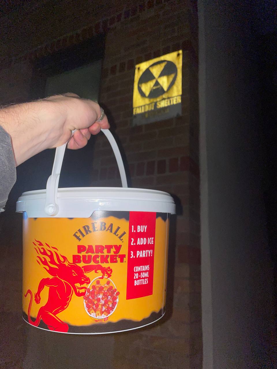 A hand is holding a large orange bucket with the label "Fireball Party Bucket" which also states it contains 20 50-mL bottles. On the bucket, there's an image of a devil or demon figure, invoking a fiery theme. There are instructions that say 1. BUY 2. ADD ICE 3. PARTY!