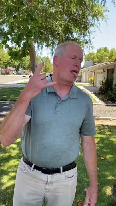 Middle-aged man, green polo shirt, white pants, outside, gesturing with hand, squinting, grass, trees, residential street.