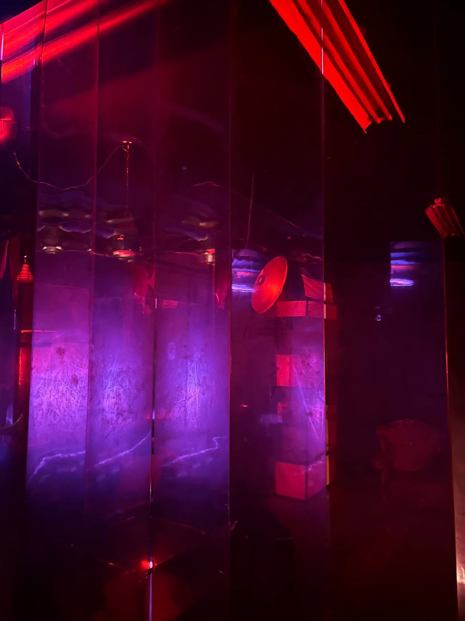 Red and purple lighting, reflective surfaces, ambiguous interior space, moody atmosphere, possibly a nightclub or a themed event space, metallic and glossy textures, obscured objects, hints of furniture, blurred motion, and an intimate or secretive vibe.