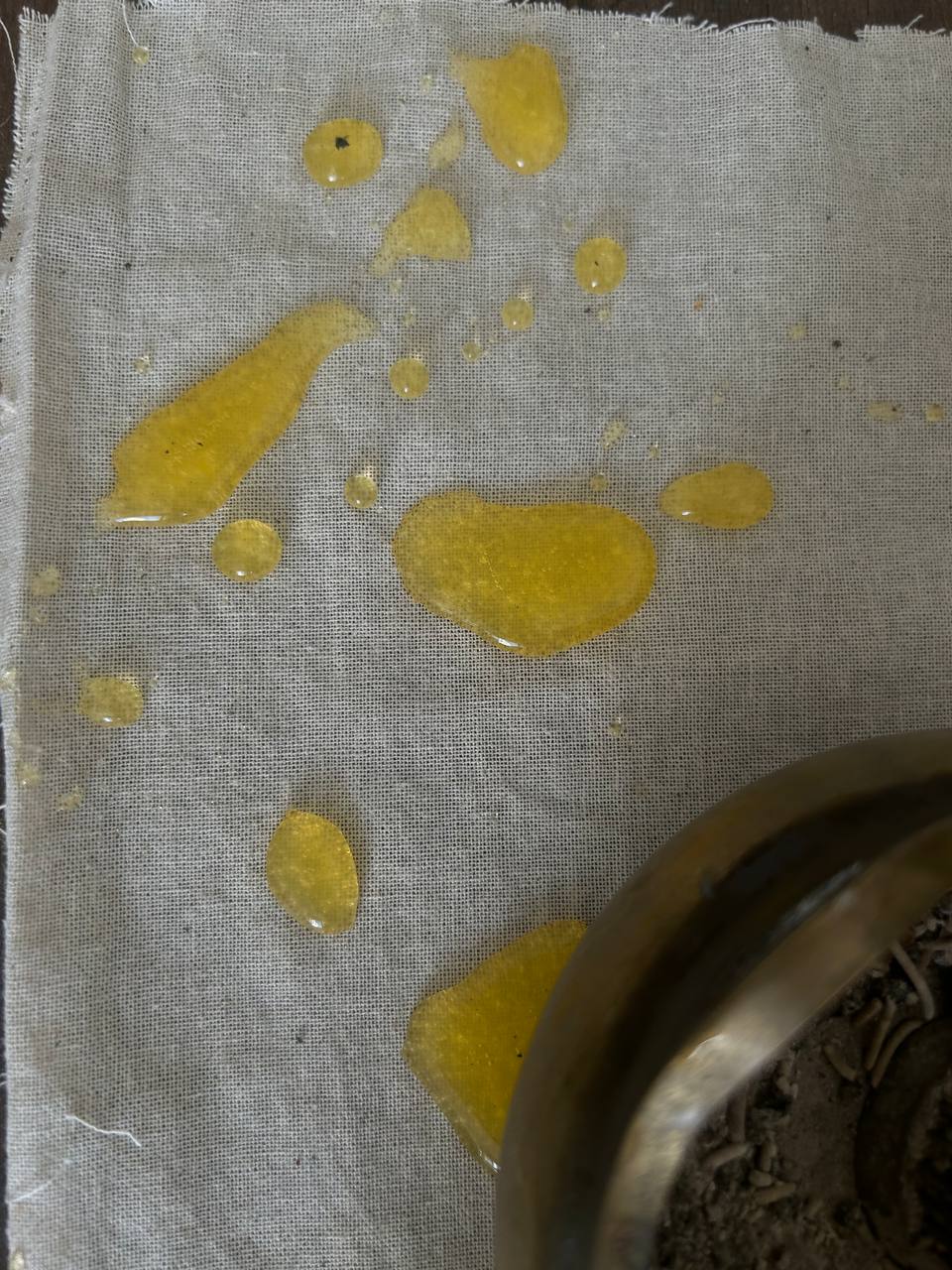 Spilled liquid, possibly oil, in various droplet sizes on a cloth. Edge of a metal or ceramic vessel with possible plant matter or dirt inside at the image's bottom right corner.