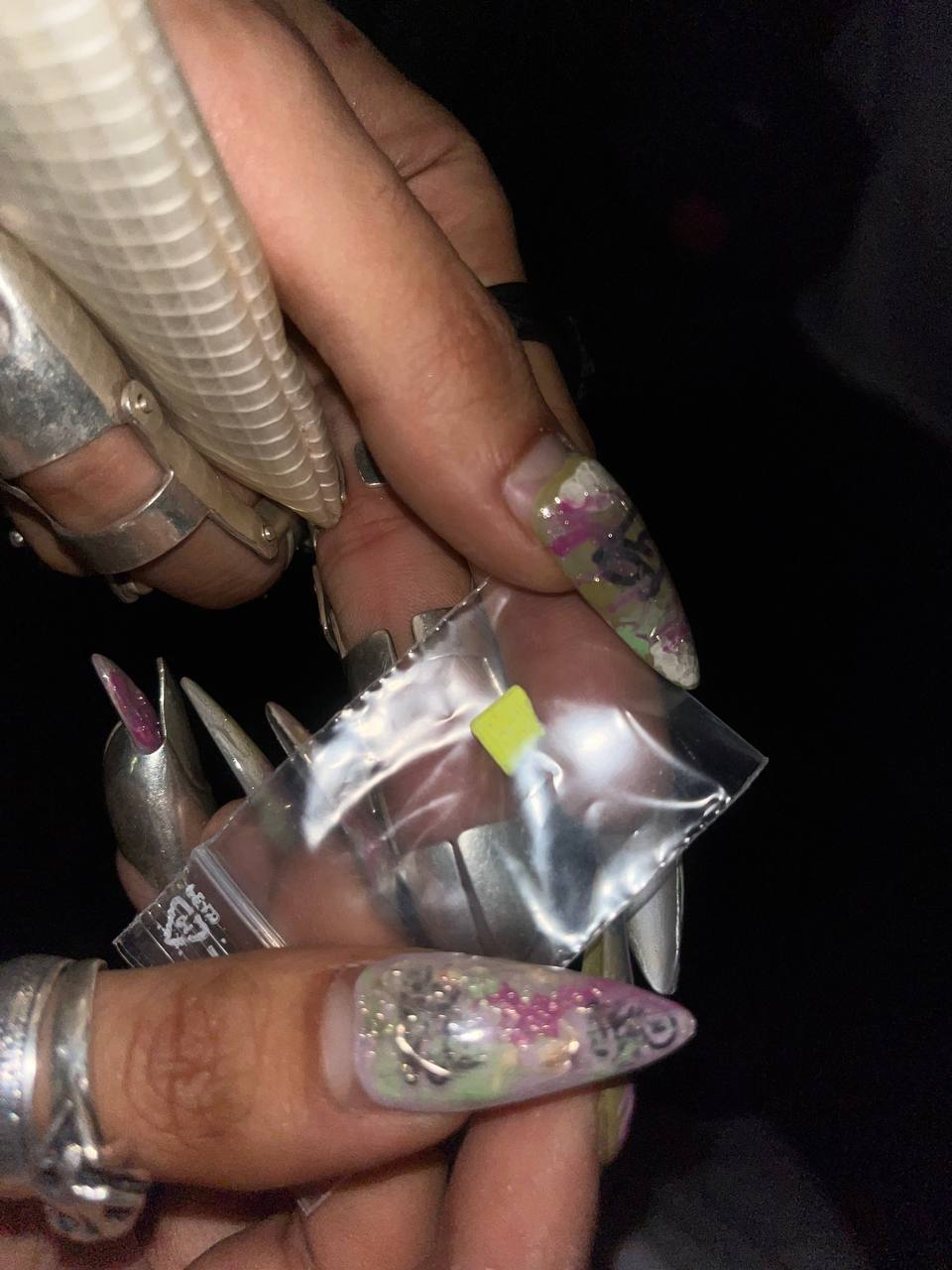 Hand holding plastic bags, elaborate fingernails with designs, silver rings, dark background.