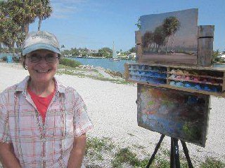 Person smiling, cap, glasses, plaid shirt, easel, colorful palette, painting, palm trees, sunny day, outdoor setting.