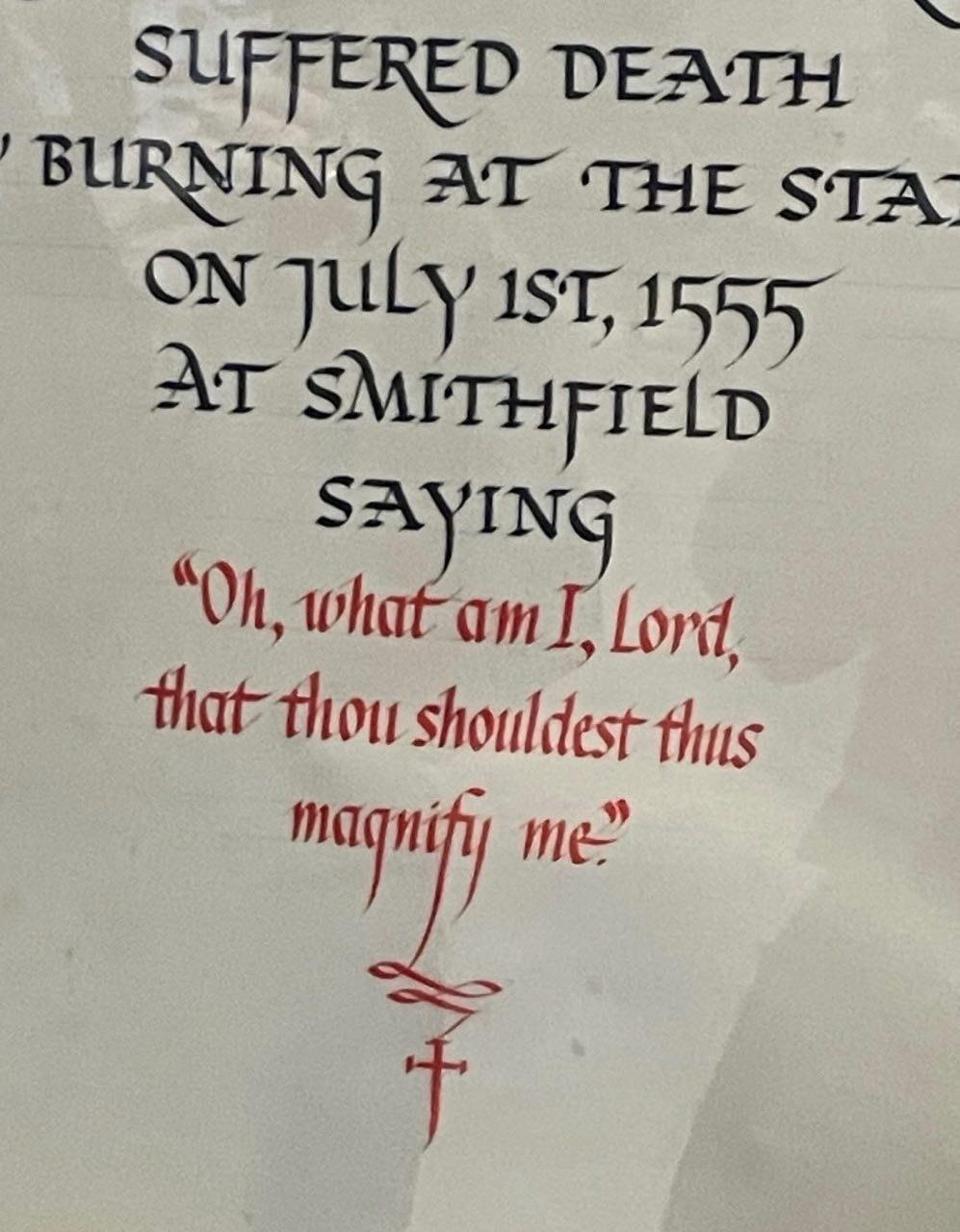 A section of text with Gothic script, apparently historical, mentioning a death by burning at the stake on July 1st, 1555, at Smithfield. A quote is given in red, "Oh, what am I, Lord, that thou shouldest thus magnify me?" Below the text is a Christian cross with a loop. The background is white and parts of letters from other words are slightly visible, indicating this is part of a larger document or display.