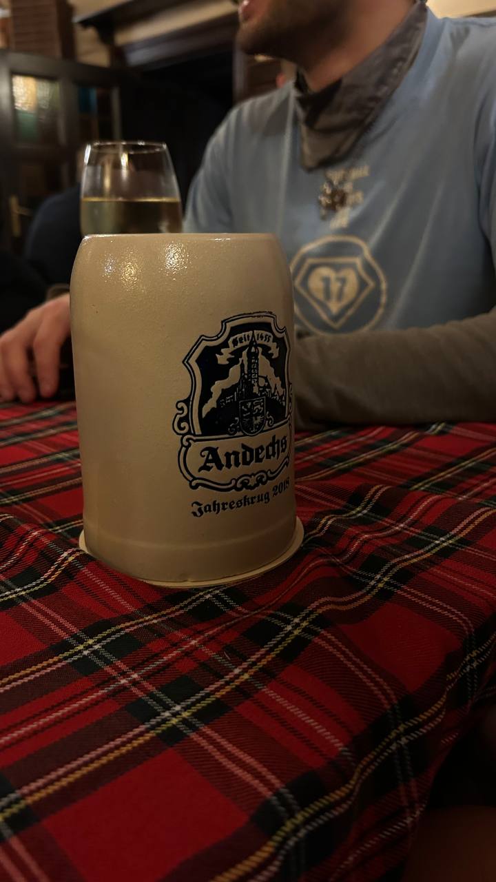 In the foreground, there's a frosted mug featuring the logo of Andechs monastery brewery established in 1455. Covered in condensation, it suggests a cold beverage is inside. On the table with a red and black tartan pattern, there is also a glass of what appears to be white wine sitting behind the mug.