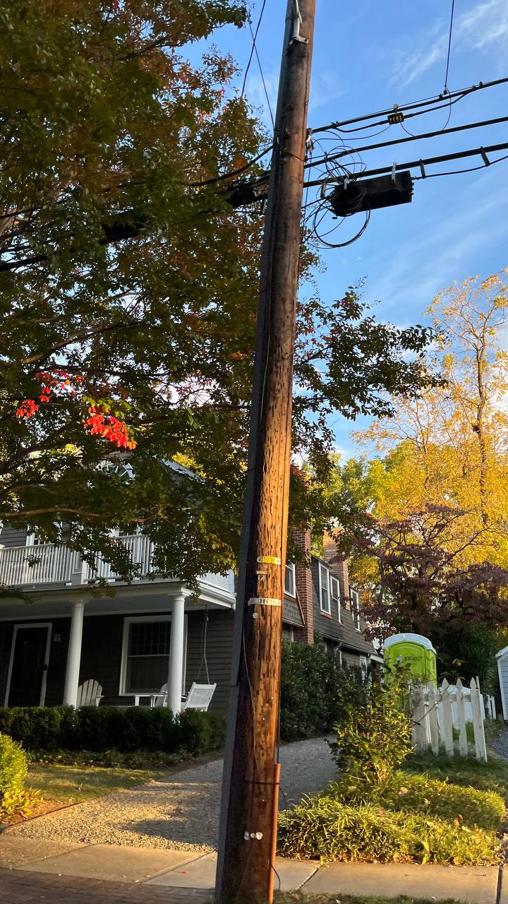 The scene includes a wooden utility pole centered closely in the foreground, equipped with electrical and communication wires, with some transformers and excess looping cables attached. In the background on is suburban housing with prominent characteristics like front porches, peaks, and a white picket fence.