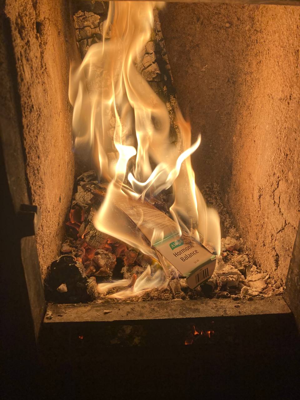 There are bright flames engulfing pieces of wood and paper within a metal or stone containment structure, which appears to be a fireplace or a stove. Some of the materials inside, specifically a carton container, can be partially seen through the flames as they burn.