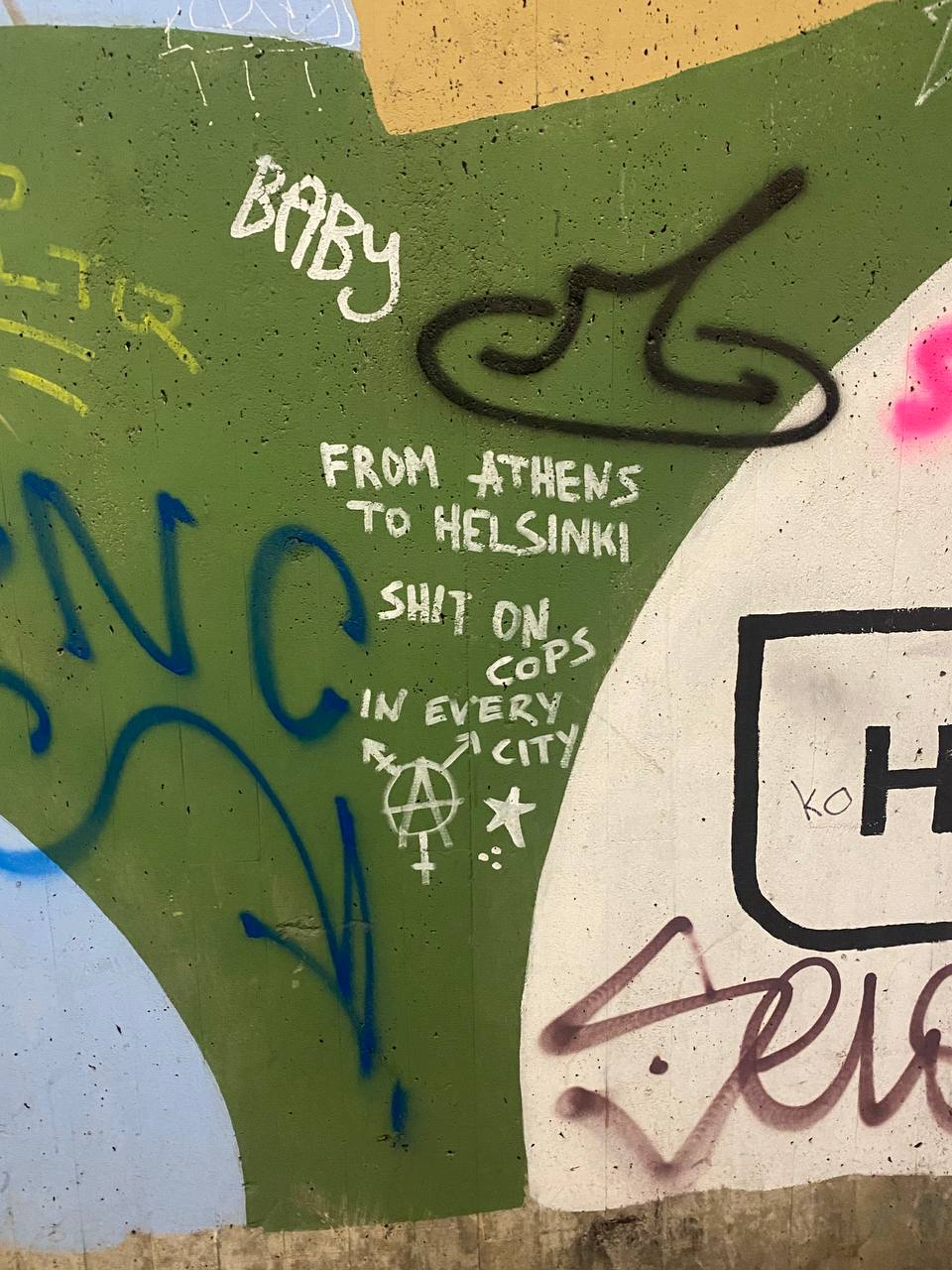 Graffiti is observed on a wall with various colors in the background, including greens and creams. The text appears to defame law enforcement with a message stating disapproval of police that extends from Athens to Helsinki.