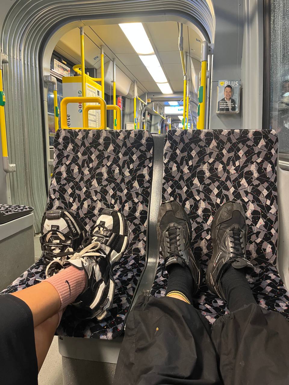 There are two pairs of feet wearing sneakers; one pair in white and black sneakers with pink socks and the other in black sneakers with black socks, both are rested on a grey and black patterned seat.