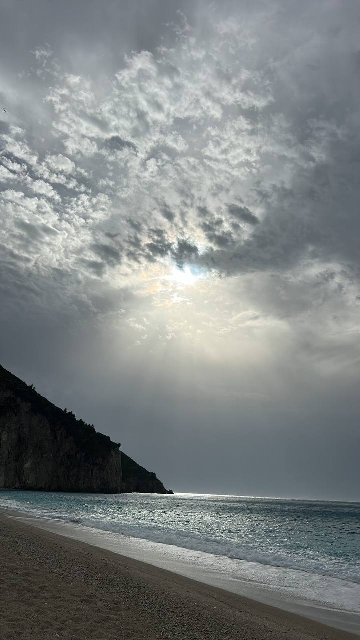 Overcast skies loom over a serene beach, where greyish clouds break just enough to allow beams of sunlight to peek through. The sea meets the shore calmly with gentle waves, and a steep cliff flanks the beach on one side, creating a rugged and dramatic contrast to the soft beach sands. The atmosphere suggests a mood of quiet introspection or a calm before a brewing storm.