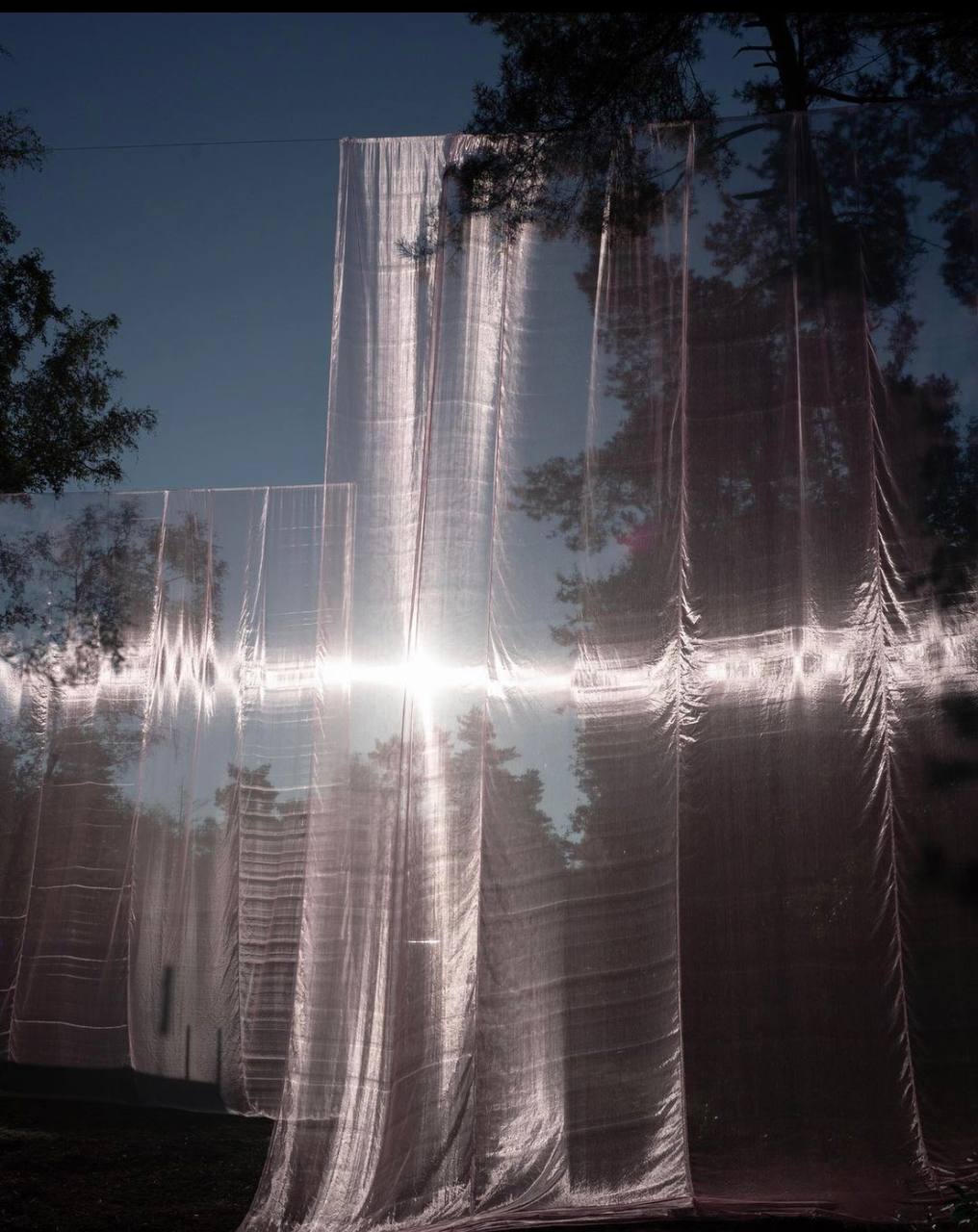 Large pieces of translucent fabric are suspended in an outdoor setting, likely hung from tree branches, rippling gently in a spacious configuration.