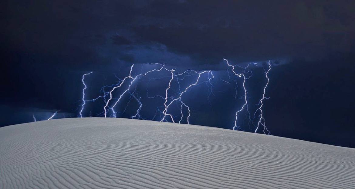 A striking natural scene with multiple lightning bolts breaking through a dark thunderstorm sky, seen from over the crest of a gently sloping sand dune. The scene shows the convergence of the powerful force of a thunderstorm contrasted with the calm, textured surface of the rippled sandy foreground.