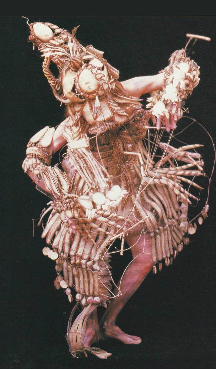 The content depicts a rhythmic and dynamic costume constructed with elements that resemble bones and skulls, mingled with intricate beading and string elements, against a dark background.