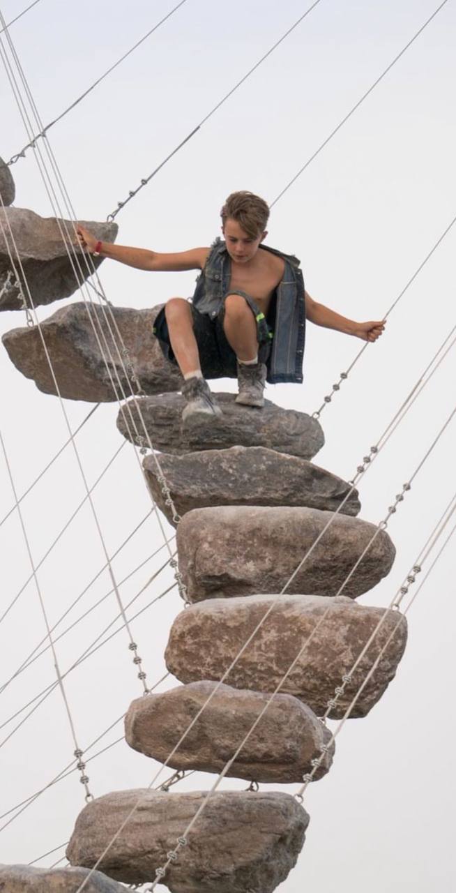 An individual appears to be climbing or navigating a vertical pathway composed of large, rough-hewn stone steps or boulders. Each of these stones is suspended by what seem to be steel cables or wires that attach the rocks and run diagonally, giving the structure stability. The background is plain, suggesting fog or an overcast sky, making the setting or location ambiguous.